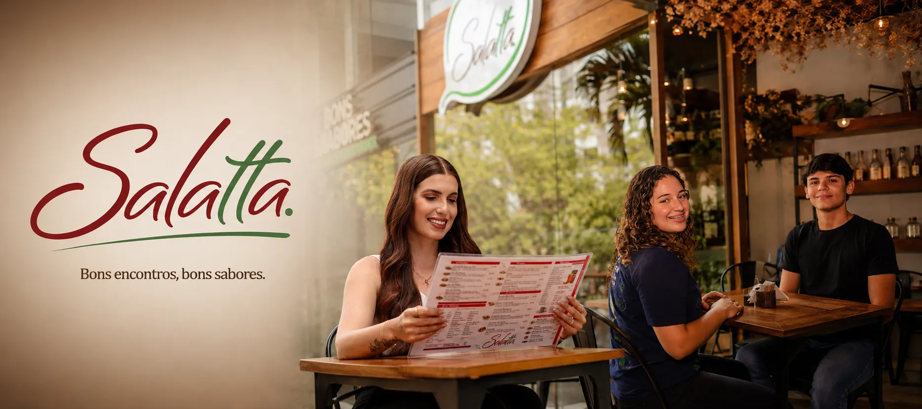 Banner do Salatta com o slogan Bons encontros, bons sabores, e clientes lendo o cardápio no restaurante em Caruaru
